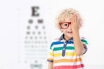 Das Bild zeigt einen Jungen mit roter Brille, der sich ein Auge zuhält. Im Hintergrund ist eine verschwommene Sehtesttafel zu sehen.